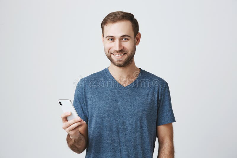 Portret van gebaard Europees mannelijk model die gelukkig camera bekijken, uitdrukkend positieve emoties, die smartphone binnen h stock afbeeldingen Portret van gebaard Europees mannelijk model die gelukkig camera bekijken, uitdrukkend positieve emoties, die smartphone binnen h stock afbeeldingen