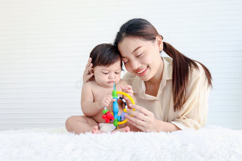 Portrait of happy crawling baby sits on fluffy white rug with mother, little cute kid girl plays with developmental toys, mom hugs her daughter child, love in family, childhood and motherhood concept. Portrait of happy crawling baby sits on fluffy white rug with mother, little cute kid girl plays with developmental toys, mom hugs her daughter child, love in family, childhood and motherhood concept