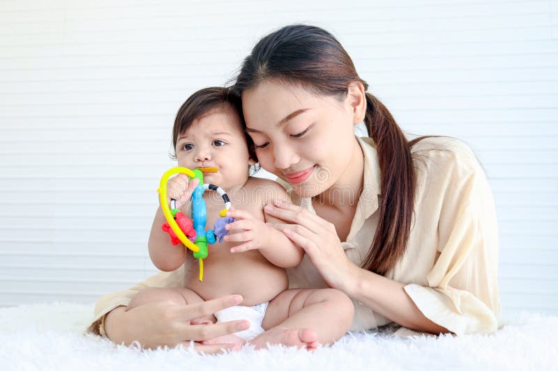 Portrait of happy crawling baby sits on fluffy white rug with mother, little cute kid girl plays with developmental toys, mom hugs her daughter child, love in family, childhood and motherhood concept. Portrait of happy crawling baby sits on fluffy white rug with mother, little cute kid girl plays with developmental toys, mom hugs her daughter child, love in family, childhood and motherhood concept