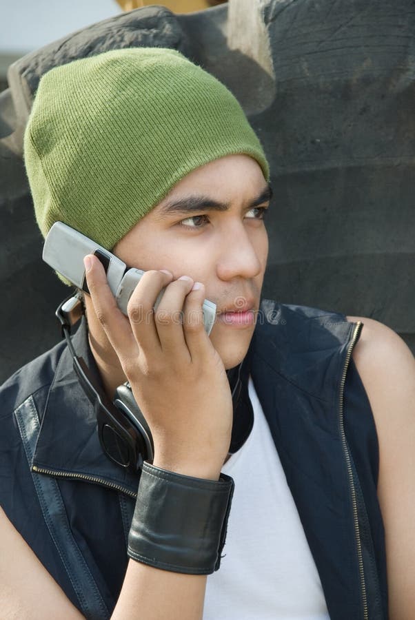 Closeup portrait of young Hispanic urban rapper dude with cap and headphone calling by cellphone against a large worn tire. Closeup portrait of young Hispanic urban rapper dude with cap and headphone calling by cellphone against a large worn tire.