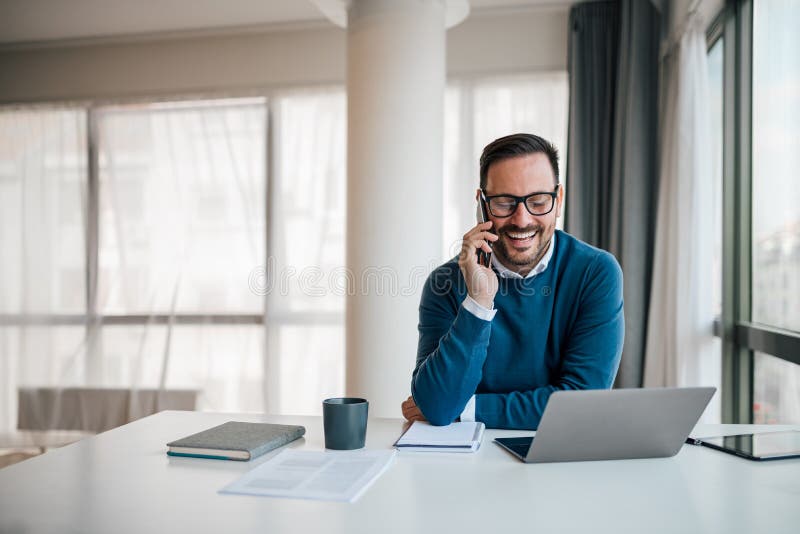 Portret van jonge glimlachende vrolijke ondernemer in een willekeurig kantoor die telefonisch belt terwijl hij werkt