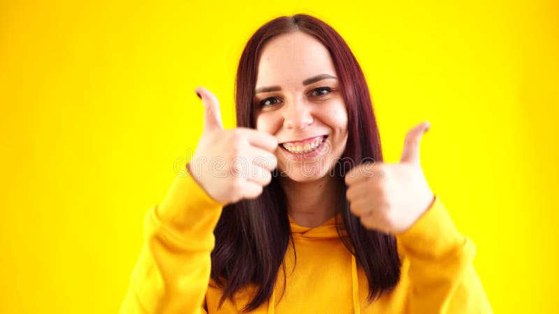 Portret van jonge vrouw die glimlacht en duimen op gele achtergrond laat zien. ' de brunette is blij dat je in de gele hoed bent '