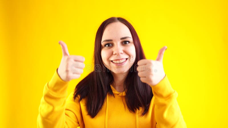 Portret van jonge vrouw die glimlacht en duimen op gele achtergrond laat zien. ' de brunette is blij dat je in de gele hoed bent '