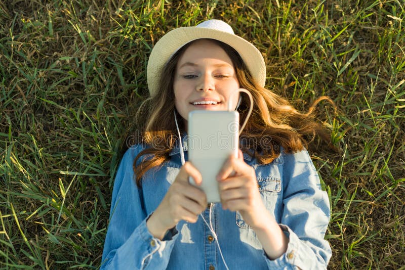 Portret van tiener 14 jaar het oude liggen op het gras Het meisje in kledingshoed, in haar hoofdtelefoons houdt een smartphone, l stock foto's Portret van tiener 14 jaar het oude liggen op het gras Het meisje in kledingshoed, in haar hoofdtelefoons houdt een smartphone, l stock foto's