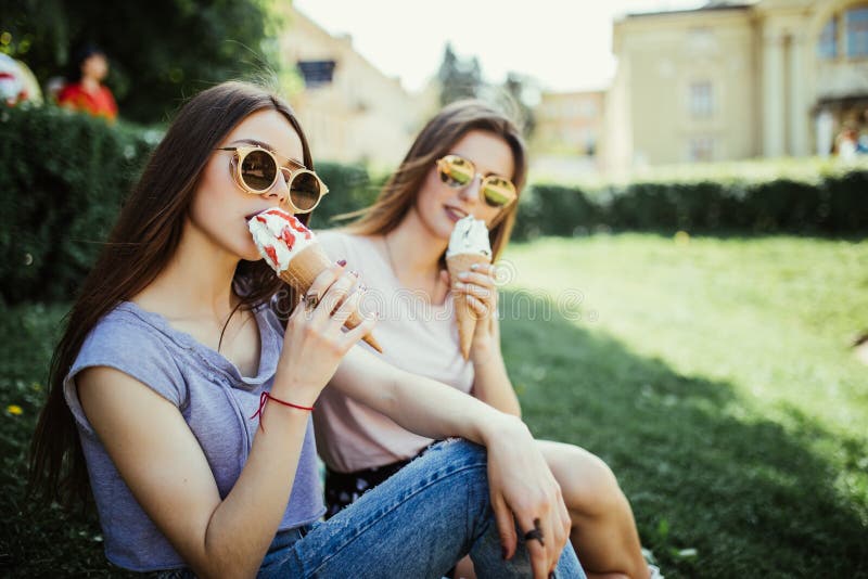 Portret van twee jonge vrouwen die op een rand zitten die roomijs in een wafelhoorn eten in de de zomerstraat royalty-vrije stock foto Portret van twee jonge vrouwen die op een rand zitten die roomijs in een wafelhoorn eten in de de zomerstraat royalty-vrije stock foto