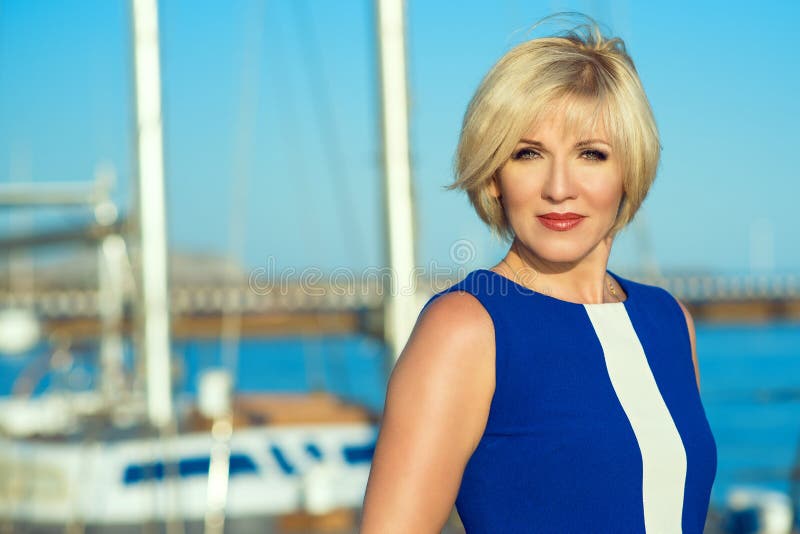 Portrait of middle-aged green-eyed gorgeous blond lady in blue dress standing on the berth. Sea background. Close up. Copy-space. Outdoor shot. Portrait of middle-aged green-eyed gorgeous blond lady in blue dress standing on the berth. Sea background. Close up. Copy-space. Outdoor shot