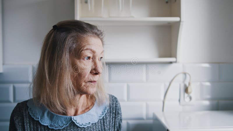 Portretul unei bătrâne cu părul gri care se uită prin geamul bucătăriei și se gândește. persoane vulnerabile singuratice stock photography
