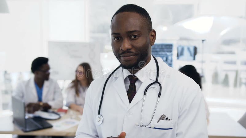 Portretul unui medic african încrezător care se uită la cameră cu o echipă de colegi pe fundal în spital. stock footage