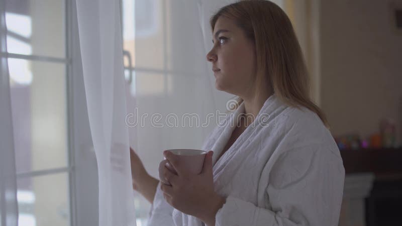 Porträt des Plusgrößenmädchens in der weißen Bademantelstellung nahe großem Fenster vom Boden bis zur Decke mit Tasse Kaffee oder