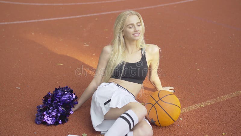 Porträt der freundlichen jungen Thin Cheerleader auf Freiengericht mit Korbball pom pom sitzt.. Lächeln blond stock footage
