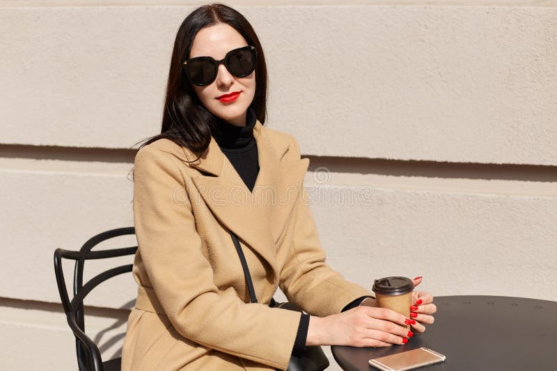 Porträt der jungen schönen brunette Frau in der stilvollen Sonnenbrille und im beige modernen Mantel sitzt bei Tisch Café im im F lizenzfreies stockbild Porträt der jungen schönen brunette Frau in der stilvollen Sonnenbrille und im beige modernen Mantel sitzt bei Tisch Café im im F lizenzfreies stockbild