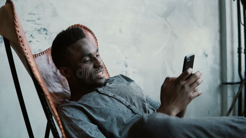 Porträt des afrikanischen Mannes sitzend im Stuhl, unter Verwendung Smartphones Hübscher Mann lächelt und betrachtet Fotos in sei lizenzfreie stockfotografie Porträt des afrikanischen Mannes sitzend im Stuhl, unter Verwendung Smartphones Hübscher Mann lächelt und betrachtet Fotos in sei lizenzfreie stockfotografie