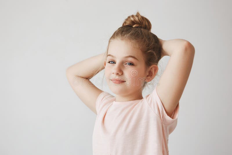 Porträt des netten sorglosen Kindes mit dem blonden Haar in der Brötchenfrisur brightfully lächelnd, Händchenhalten hinter Kopf