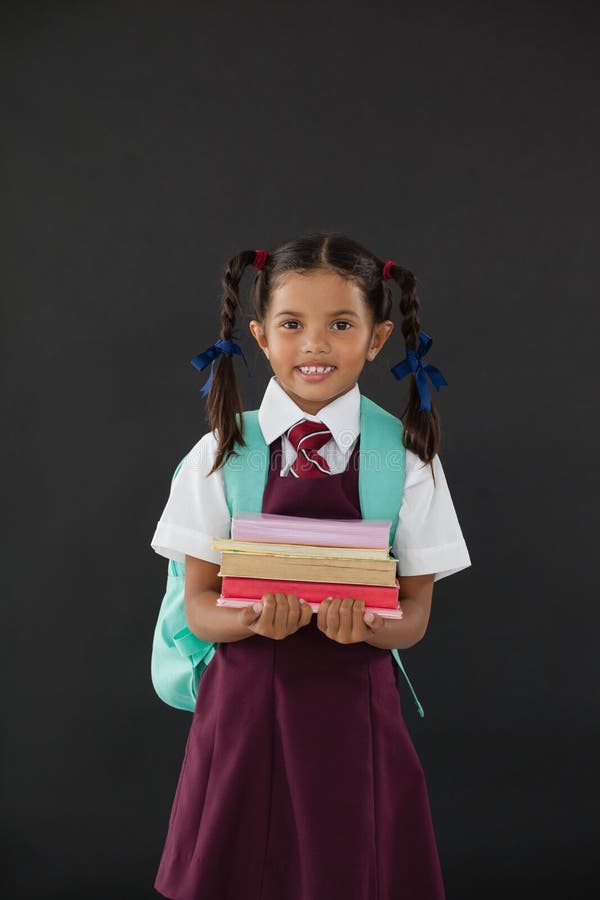 Porträt des Schulmädchens in der Schuluniform, die Bücher gegen Tafel hält