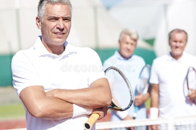 Porträt des überzeugten reifen Mannes, der Tennisschläger bei der Stellung mit den Armen gekreuzt gegen Freunde auf Gericht währe stockbild Porträt des überzeugten reifen Mannes, der Tennisschläger bei der Stellung mit den Armen gekreuzt gegen Freunde auf Gericht währe stockbild