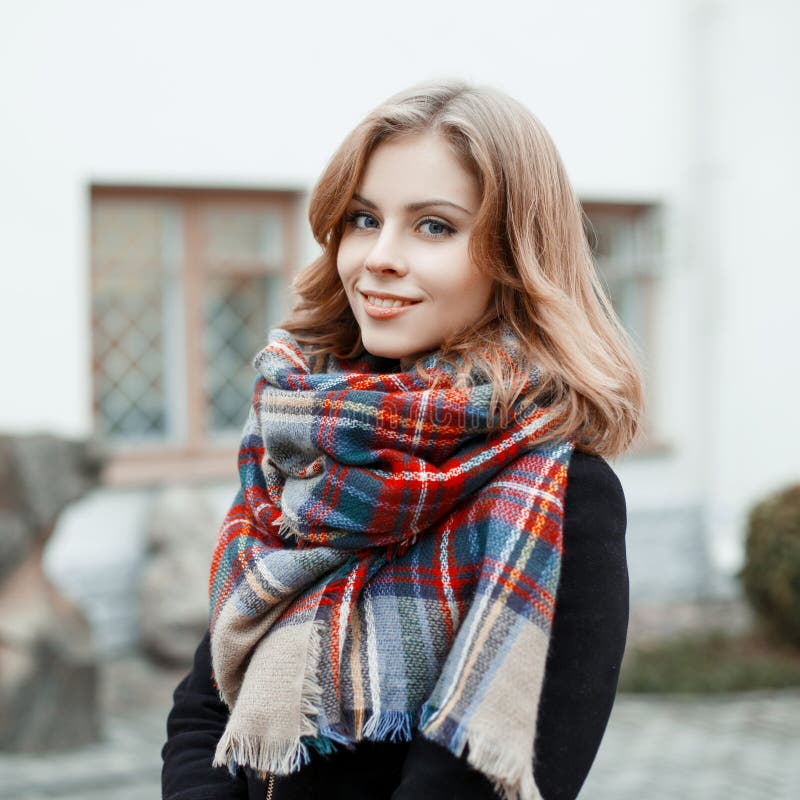 Portrait of an attractive woman with blond hair with a wonderful smile in a black stylish coat with a woolen checkered warm scarf on the background of a gray historic building. Lovely girl smiling. Portrait of an attractive woman with blond hair with a wonderful smile in a black stylish coat with a woolen checkered warm scarf on the background of a gray historic building. Lovely girl smiling