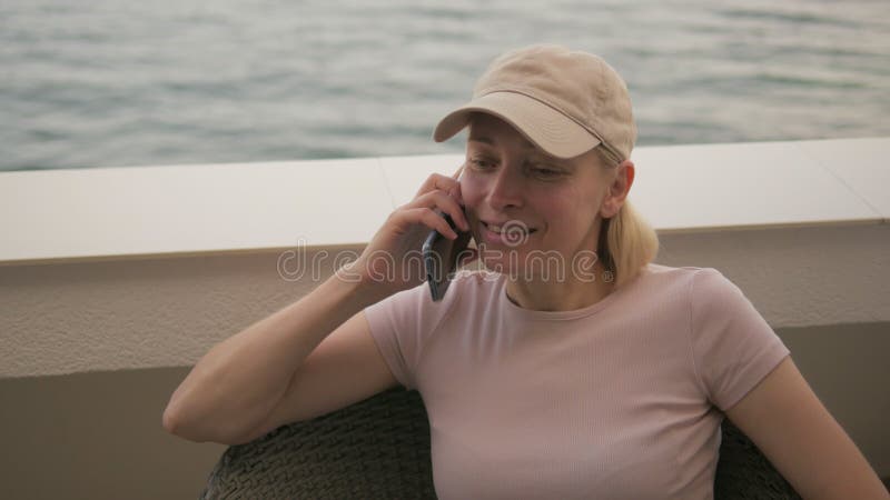 Porträt einer Frau mit Baseballkappe, die auf dem Telefon sitzt und auf dem Balkon eines Hotelzimmers mit Blick auf das Meer sitzt