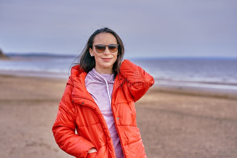 Porträt einer jungen Brunette-Frau in Sonnenbrille und roter Jacke Frau auf einem Spaziergang entlang der Sandküste eines großen stockfotografie