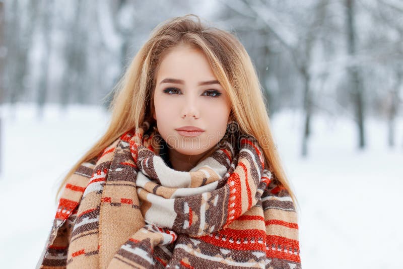 Portrait of a pretty young woman with brown eyes with beautiful makeup with long blond hair in a woolen vintage warm scarf in a snowy park. Cute girl on a walk. Portrait of a pretty young woman with brown eyes with beautiful makeup with long blond hair in a woolen vintage warm scarf in a snowy park. Cute girl on a walk