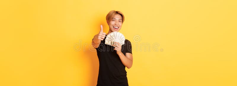 Porträt eines ansehnlichen, zuversichtlich lächelnden asiatischen Mannes, der Daumen zeigt und Geld hält, garantiert etwas Standha lizenzfreies stockbild