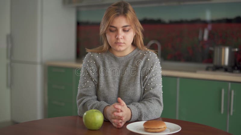 Porträt eines fetten Mädchens, das eine Wahl zwischen der gesunden und ungesunden Nahrung sitzt am Tisch in der Küche trifft Das