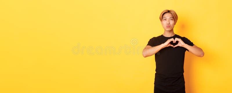 Porträt eines hübschen blonden asiatischen Mannes, der eine Herzgeste und einen gelben Hintergrund mit Lächeln zeigt stockbild