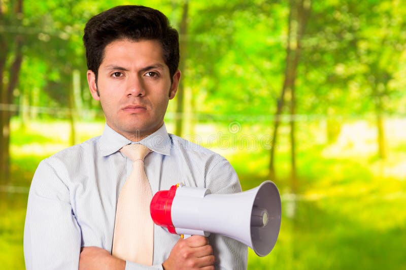 Porträt eines hübschen wütenden Mannes, der in seinem Hand ein Megaphon mit seinen Armen gekreuzt, in einem unscharfen grünen Hin lizenzfreie stockfotos Porträt eines hübschen wütenden Mannes, der in seinem Hand ein Megaphon mit seinen Armen gekreuzt, in einem unscharfen grünen Hin lizenzfreie stockfotos