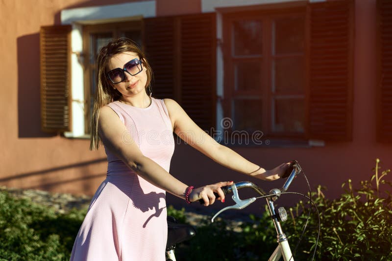 Porträt eines jungen, attraktiven Mädchens in den Gläsern und des rosa Kleides auf einem Fahrrad lizenzfreies stockbild Porträt eines jungen, attraktiven Mädchens in den Gläsern und des rosa Kleides auf einem Fahrrad lizenzfreies stockbild