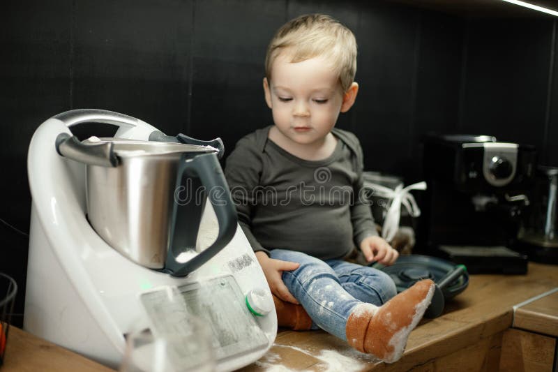 Portrait of cherubic little boy baby toddler with short fair hair sitting on table near kneader cooking machine in black kitchen at home, looking at floury leg. Childhood, cooking, baking, pastry. Portrait of cherubic little boy baby toddler with short fair hair sitting on table near kneader cooking machine in black kitchen at home, looking at floury leg. Childhood, cooking, baking, pastry.