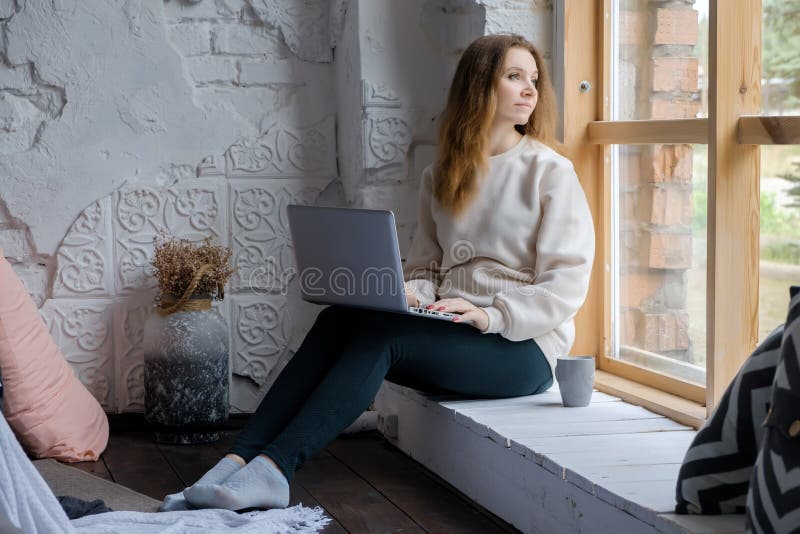 Porträt eines schönen jungen Mädchens, das auf einem Fensterbrett morgens mit einem Laptopbuch auf ihren Knien sitzt und heraus s lizenzfreies stockfoto Porträt eines schönen jungen Mädchens, das auf einem Fensterbrett morgens mit einem Laptopbuch auf ihren Knien sitzt und heraus s lizenzfreies stockfoto