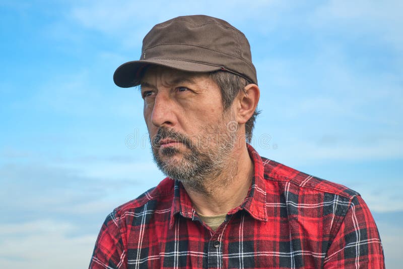 Portrait of serious confident male farmer with brown cap and red laid shirt against blue sky, selective focus. Portrait of serious confident male farmer with brown cap and red laid shirt against blue sky, selective focus