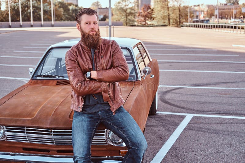 Portrait of a stylish bearded male in old-fashioned clothes sits on the hood retro car in the city parking. Country style concept. Portrait of a stylish bearded male in old-fashioned clothes sits on the hood retro car in the city parking. Country style concept.