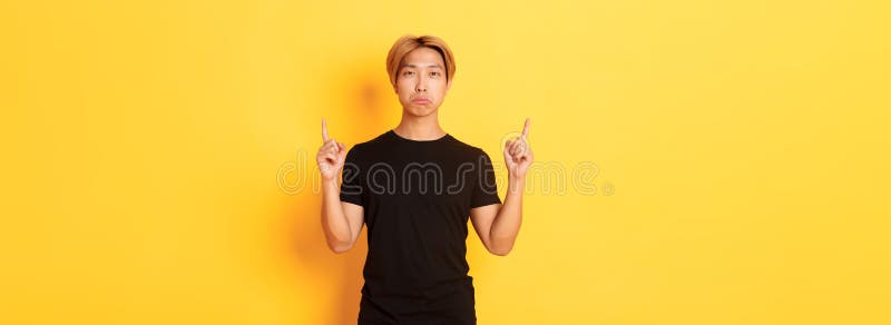 Porträt eines trostlosen dunklen asiatischen Mannes, der enttäuscht aussieht, wenn er die Finger auf gelben Hintergrund zeigt stockbild