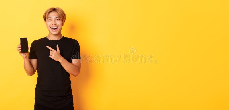 Porträt eines zufriedenen lächelnden Asiaten mit blonden Haaren, amüsant aussehen und mit dem Finger auf Smartphone-Bildschirm zei stockbild