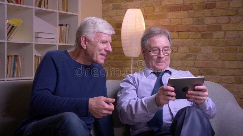Porträt von den älteren männlichen Freunden, die zusammen auf dem Sofa aufpasst in Tablette und froh sich bespricht sitzen