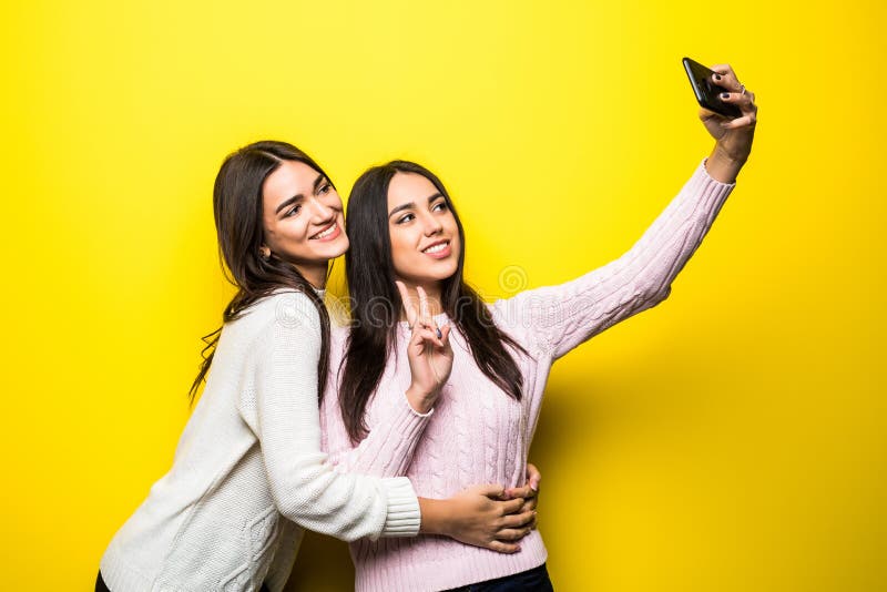 Portrait of two lovely girls dressed in sweaters standing and taking a selfie over yellow. Portrait of two lovely girls dressed in sweaters standing and taking a selfie over yellow