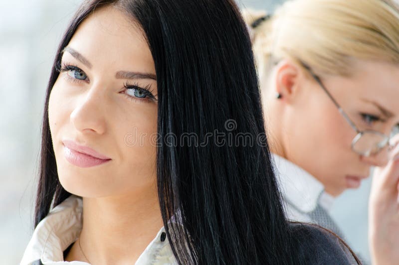 Portrait of two beautiful young women with blue eyes brunette & blond co-workers near office window at daytime. Portrait of two beautiful young women with blue eyes brunette & blond co-workers near office window at daytime