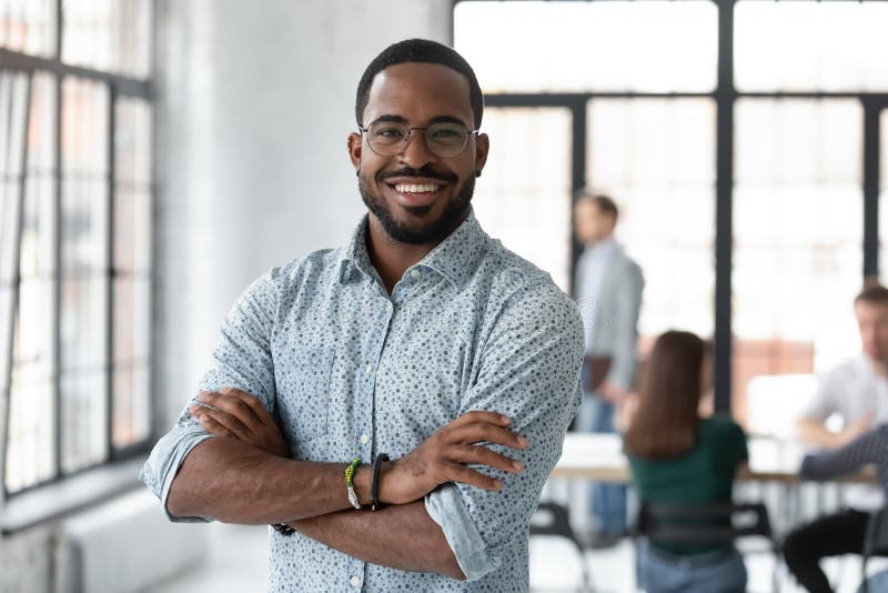 Portrait of happy African American small business owner posing with hands folded. Millennial black male team leader smiling, looking at camera, employees working in modern office behind. Head shot. Portrait of happy African American small business owner posing with hands folded. Millennial black male team leader smiling, looking at camera, employees working in modern office behind. Head shot