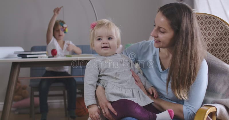 Porträtt av en glad blond flicka som sitter med mor på en roterande stol och leende Hennes äldre bror leker med leksak