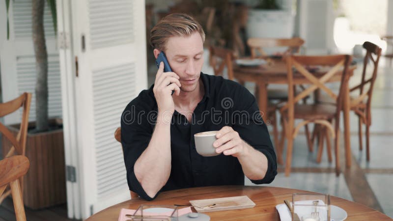 Porträtt av en stilig man som dricker kaffe i modern kafé och pratar på telefon lager videofilmer