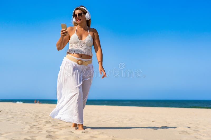 Porträtt av en vacker medelålders kvinna som lyssnar på musik i hörlurar som håller smartphone på sandstranden. arkivfoton