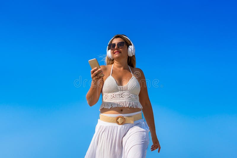 Porträtt av en vacker medelålders kvinna som lyssnar på musik i hörlurar som håller smartphone på sandstranden. arkivfoton