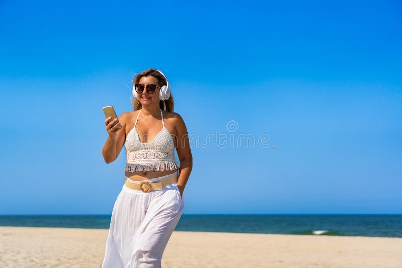 Porträtt av en vacker medelålders kvinna som lyssnar på musik i hörlurar som håller smartphone på sandstranden. royaltyfria bilder