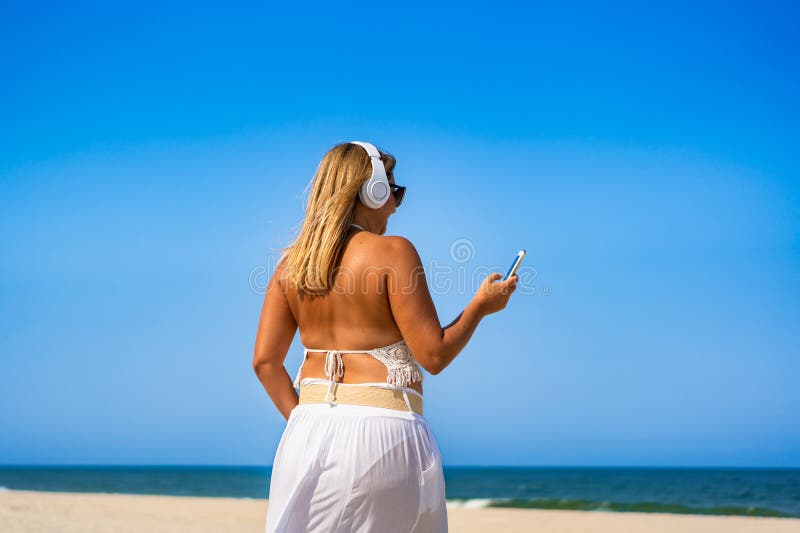 Porträtt av en vacker medelålders kvinna som lyssnar på musik i hörlurar som håller smartphone på sandstranden. royaltyfria foton