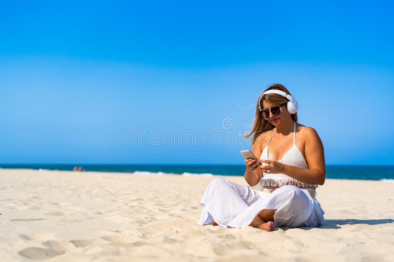 Porträtt av en vacker medelålders kvinna som lyssnar på musik i hörlurar som håller smartphone sittande på sandstranden arkivbild