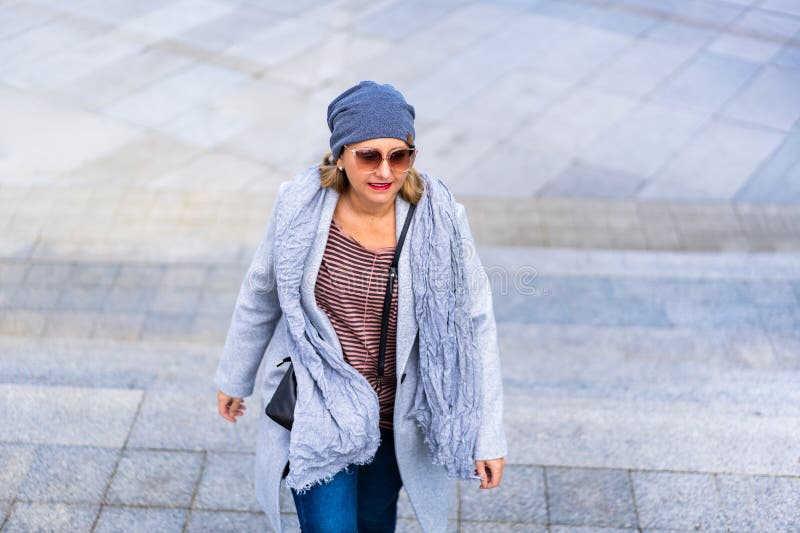 Porträtt av en vuxen blondinkvinna i en grå sjökort jeans vinterhatt och solglasögon som går upp i trappan arkivbilder
