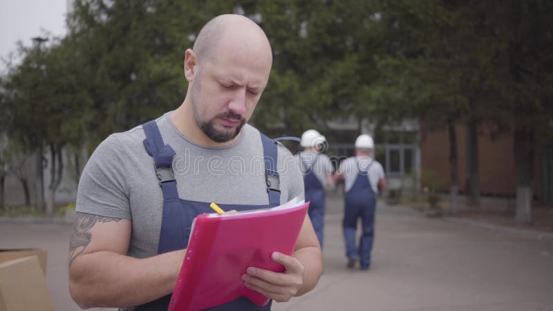 Porträtt av en vuxen skäggig kaukasisk man som skriver på fraktsedeln som två arbetare som går iväg med paket Arbeta i