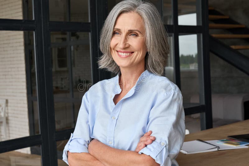 Smiling adult middle aged business woman standing at table posing in home office, arms crossed. Portrait of gray haired happy professional ceo or financial director woman in corporate photoshoot. Smiling adult middle aged business woman standing at table posing in home office, arms crossed. Portrait of gray haired happy professional ceo or financial director woman in corporate photoshoot.