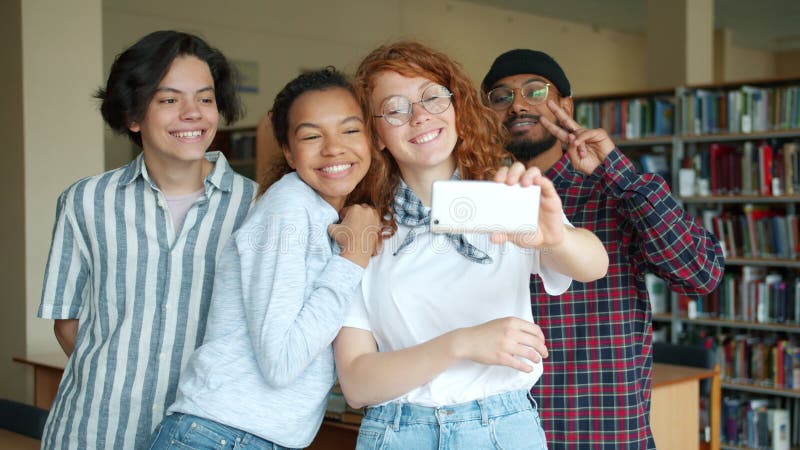 Porträtt av lyckliga studenter som tar selfie i biblioteket med smarttelefonkamera arkivfilmer