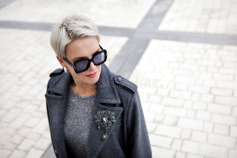 Portrait of fashionable mature woman with short blonde hair wearing stylish gray wool coat with lapels and shoulder straps, accessories brooch and sunglasses. Portrait of fashionable mature woman with short blonde hair wearing stylish gray wool coat with lapels and shoulder straps, accessories brooch and sunglasses.